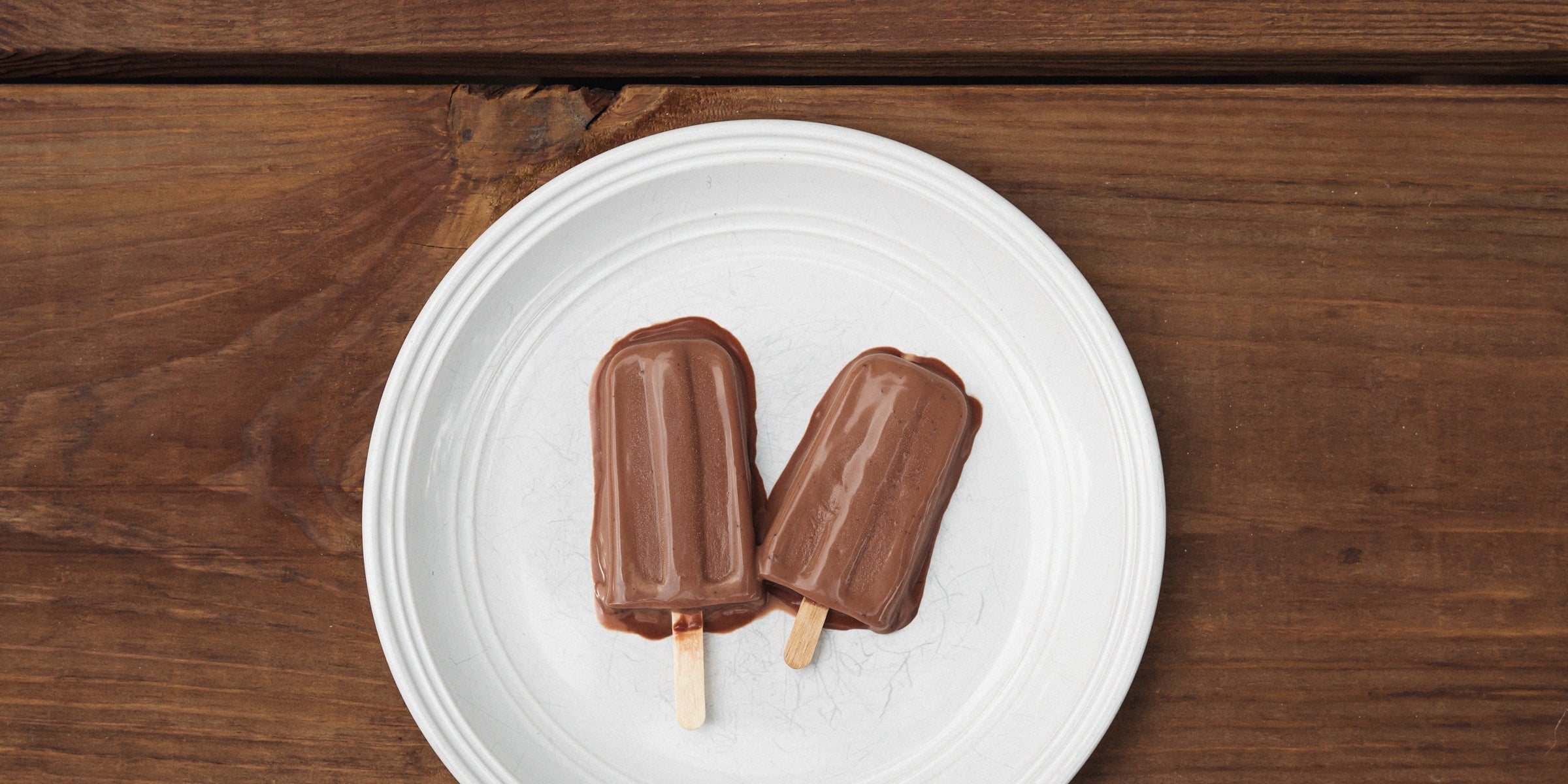 Two chocolate popsicles laid together on a white porcelain plate sitting on a picnic table