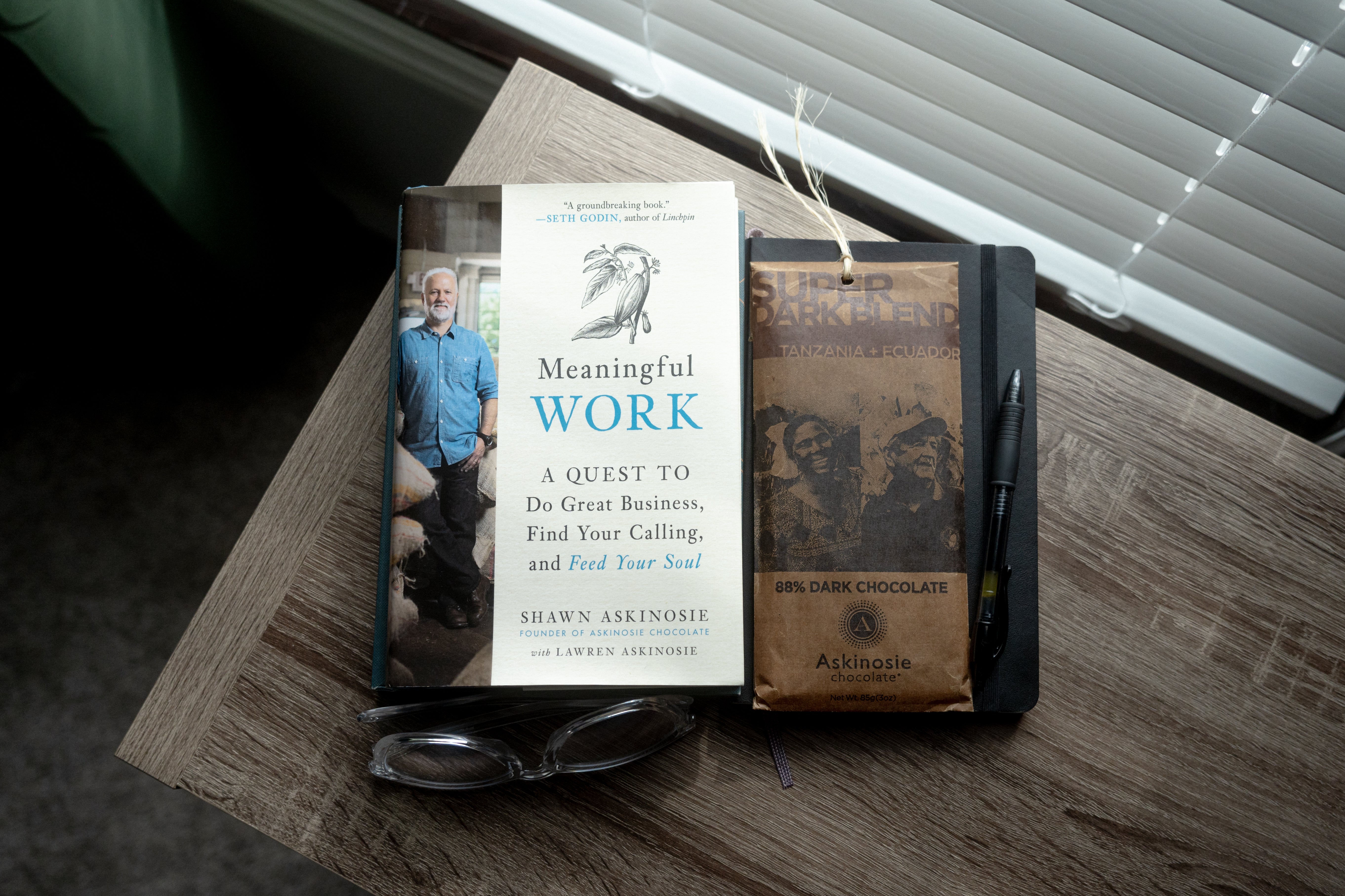 Overhead image of Meaningful Work by Shawn Askinosie with a packaged chocolate bar, glasses and a notebook stacked nearby.